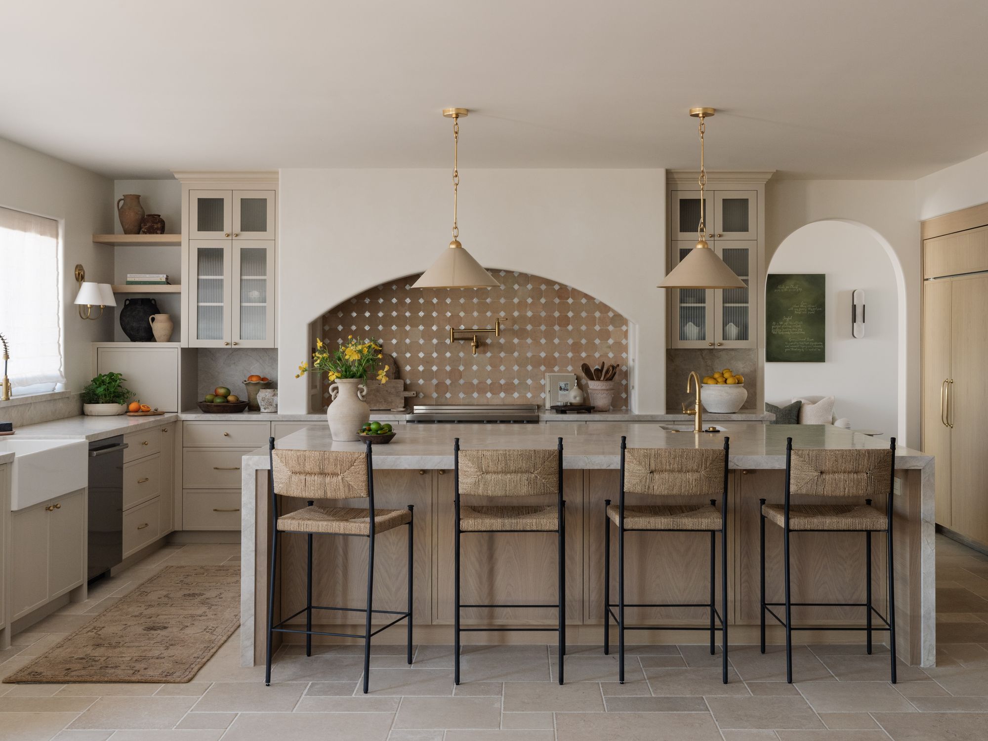A warm family kitchen with a long limestone island, woven barstools, brass lighting, and a gathering space