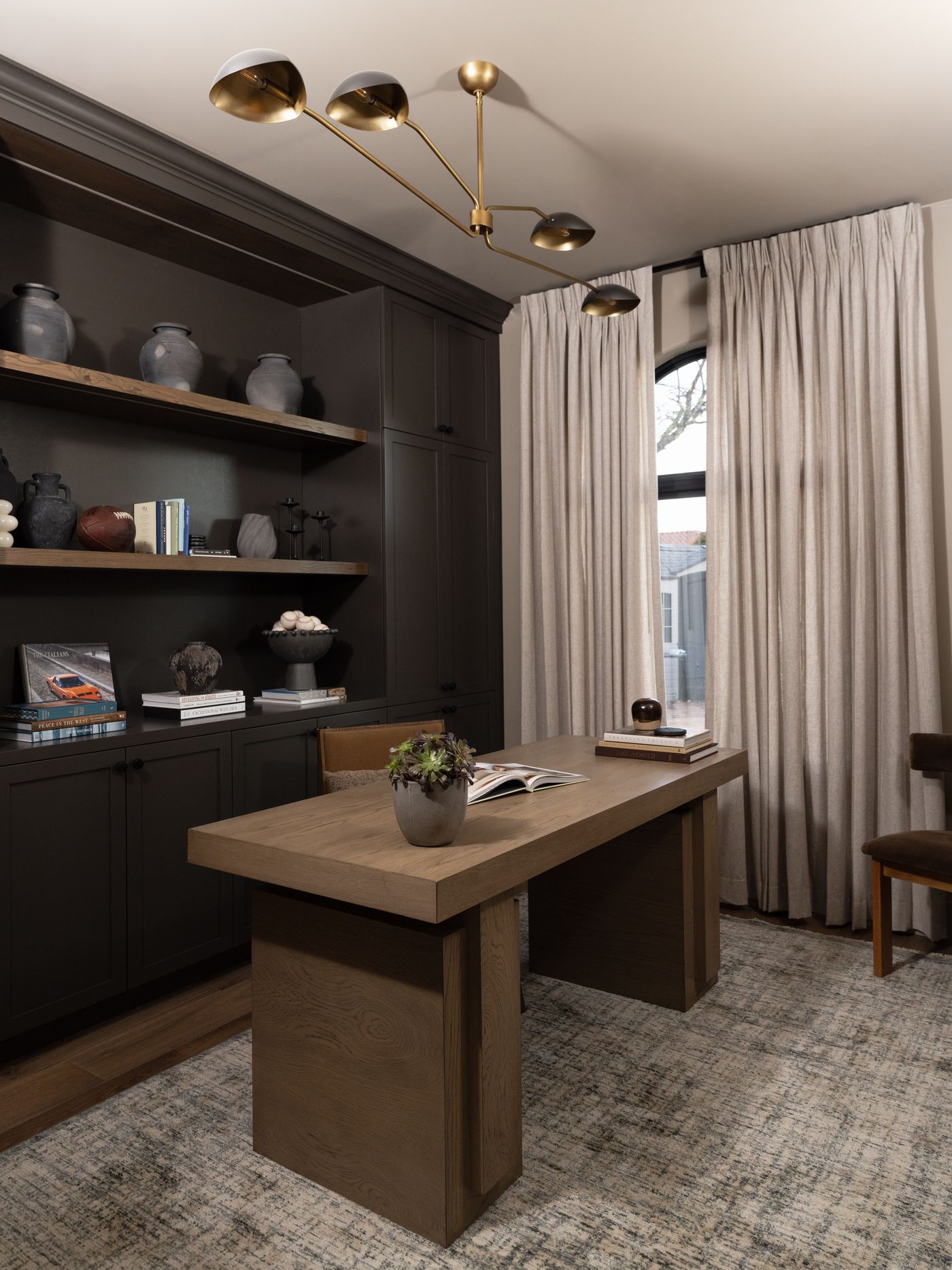 A quiet home office with dark built-in shelving, a solid oak desk, warm layered lighting, and books